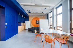 Kolorowe biuro w niebiesko-pomarańczowej tonacji, wyposażone w wygodne krzesła i funkcjonalne stoły.