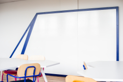 Klasa z białymi ścianami, niebieskimi i różowymi krzesłami oraz tablicą na ścianie.